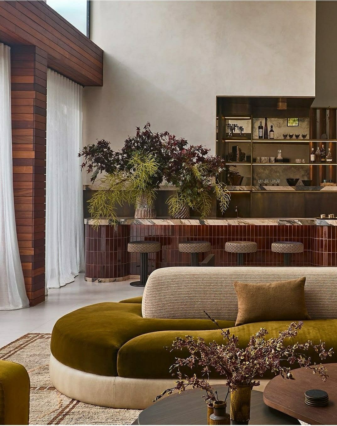 Sage green and red interior with backlit arch bottle niches, bar stools, and sculptural branches